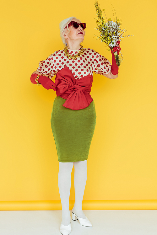 Fashionable senior woman in sunglasses holding bouquet of wildflowers on white surface on yellow background