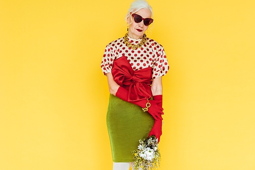 Beautiful elderly woman in sunglasses holding bouquet of wildflowers isolated on yellow