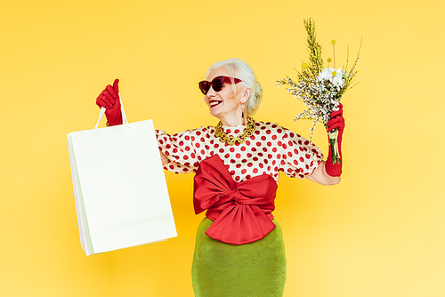 Fashionable senior woman smiling while holding wildflowers and shopping bags on yellow background