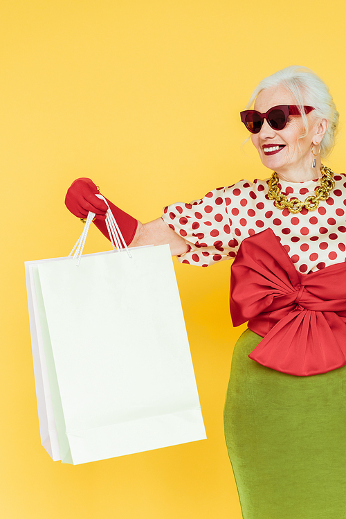 Stylish elderly woman smiling while holding shopping bags isolated on yellow