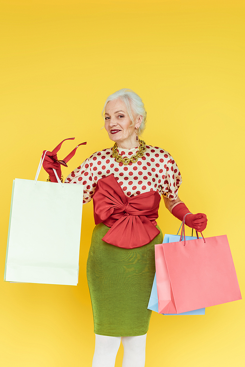 Stylish senior woman smiling at camera while holding shopping bags and sunglasses isolated on yellow