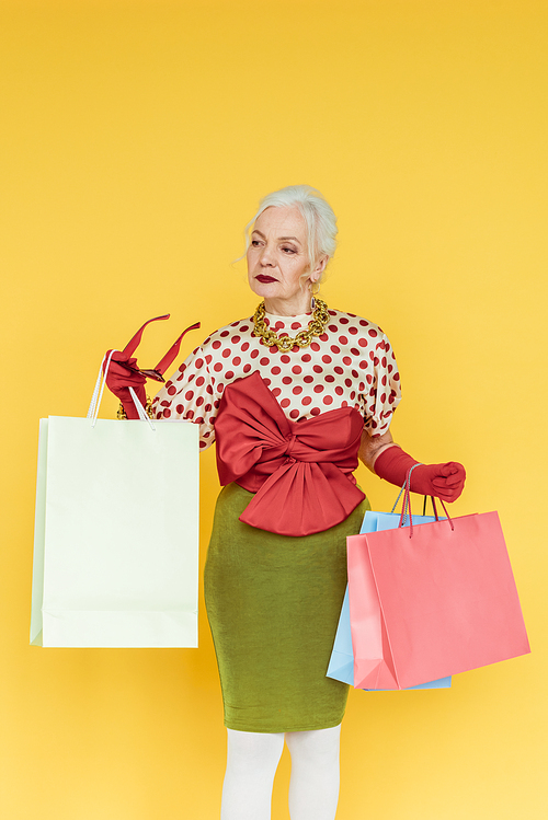 Fashionable senior woman holding shopping bags and sunglasses isolated on yellow