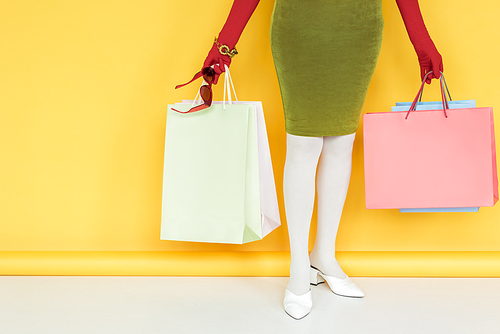 Cropped view of stylish senior woman holding sunglasses and shopping bags on white surface on yellow background