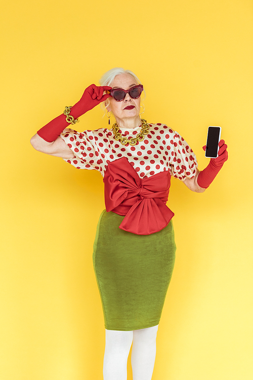 Stylish senior woman in sunglasses holding smartphone on yellow background