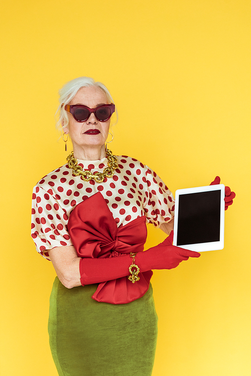 Stylish senior woman holding digital tablet isolated on yellow