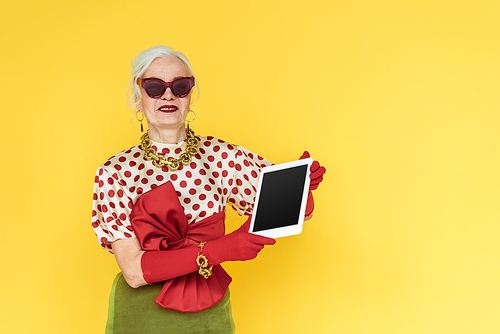 Fashionable senior woman smiling while holding digital tablet isolated on yellow