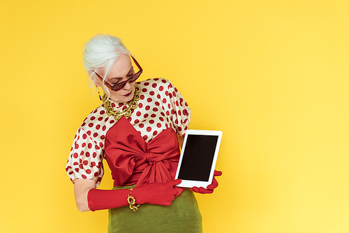 Fashionable elderly woman holding digital tablet with blank screen isolated on yellow