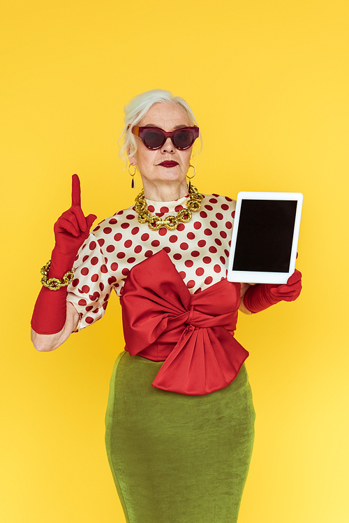 Fashionable senior woman in sunglasses holding digital tablet and pointing with finger isolated on yellow