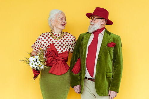 Stylish senior couple with bouquet of wildflowers smiling at each other on yellow background