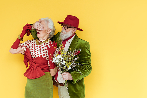 Stylish senior man covering eyes to woman and holding bouquet of wildflowers on yellow background