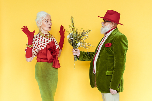 Stylish senior man giving wildflowers to surprised woman on yellow background
