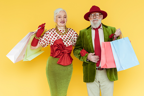 Stylish senior couple with shopping bags smiling at camera on yellow background