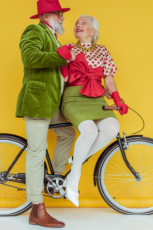 Fashionable senior couple smiling at each other on bicycle on white surface on yellow background