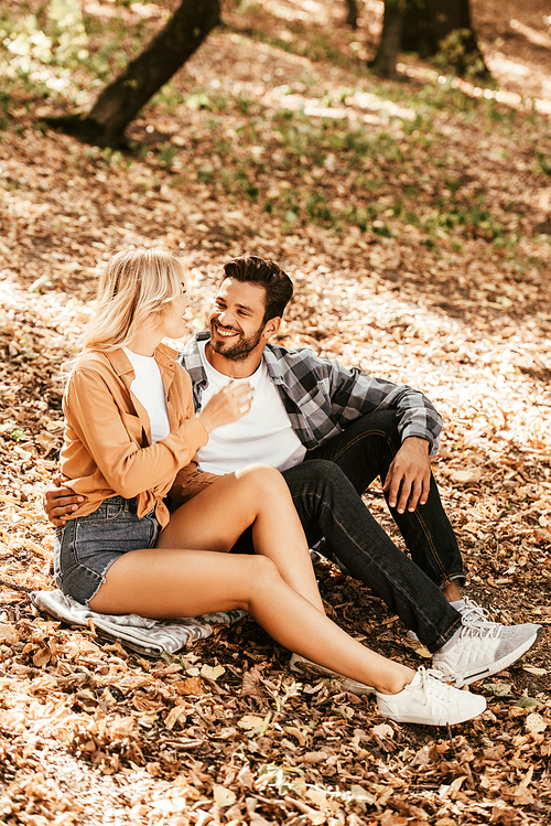 happy young couple looking at each other while sitting on fall foliage in park
