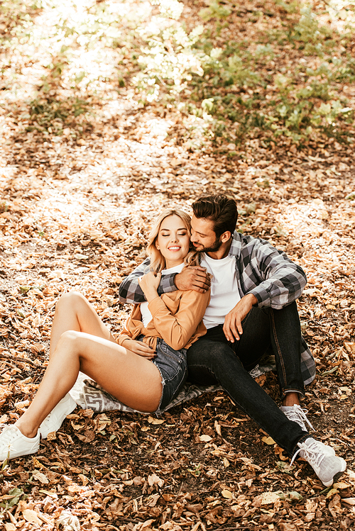 happy man embracing cheerful girlfriend while sitting on fall foliage in park
