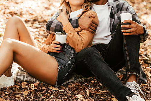 cropped view of young couple holding thermo cups while sitting on fall foliage