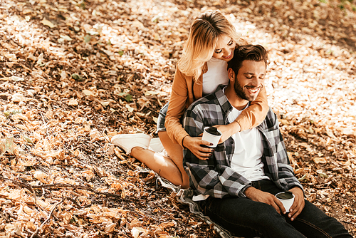 happy girl embracing cheerful boyfriend while sitting on fall foliage in park