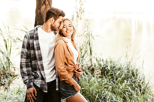 young man embracing cheerful girlfriend while standing near lake in park