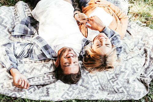 overhead view of cheerful young couple smiling while lying on blanket in park