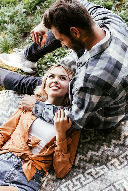 overhead view of young man sitting near smiling girlfriend lying on blanket in park