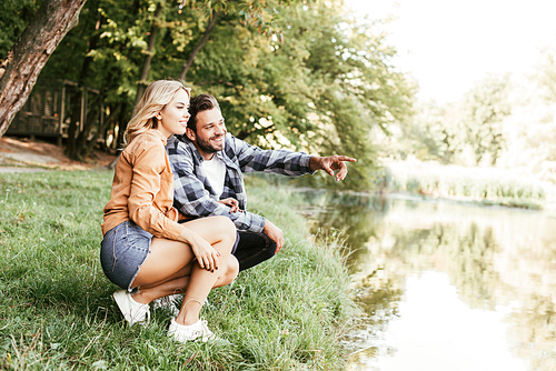 cheerful man pointing with finger while squatting near lake with girlfriend