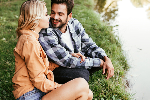 happy young couple looking at each other while squatting near lake in park