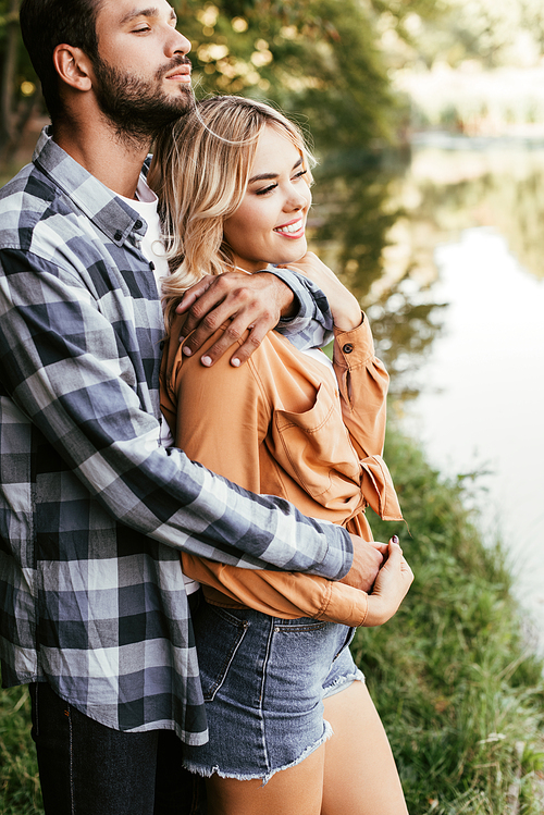 handsome man embracing smiling girlfriend while standing near lake in park