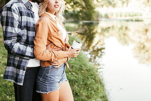 cropped view of young man embracing smiling girlfriend while standing near lake in park