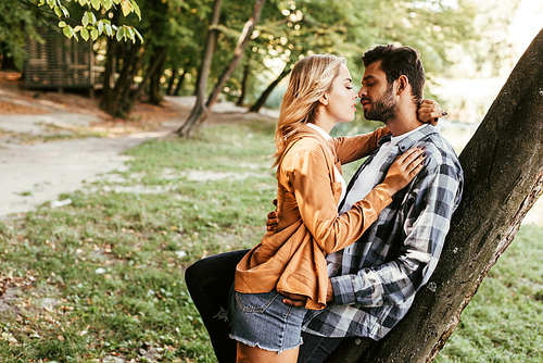 attractive young woman kissing boyfriend while standing tree trunk in park