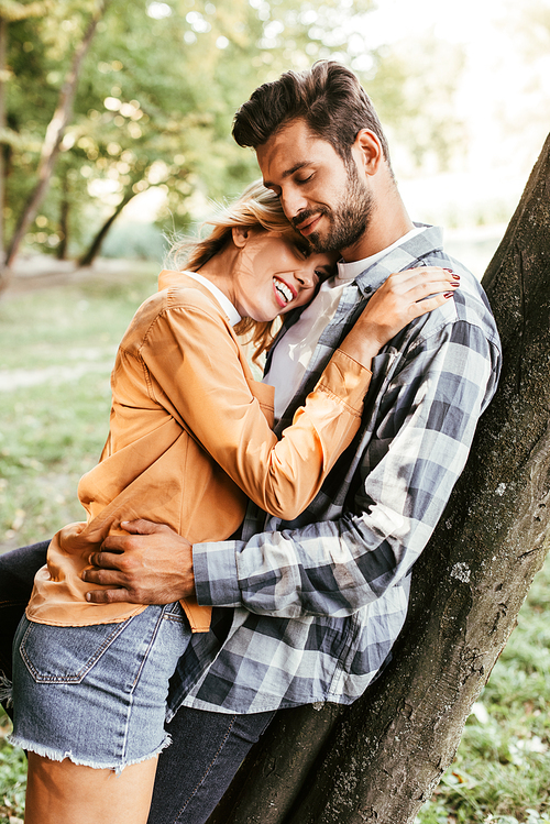 attractive young woman embracing boyfriend while standing near tree trunk in park