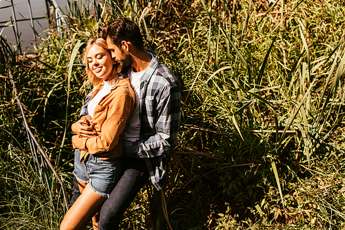 handsome young man embracing happy girlfriend in thicket of sedge near lake