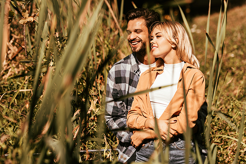 selective focus of happy young man embracing smiling girlfriend in thicket of sedge
