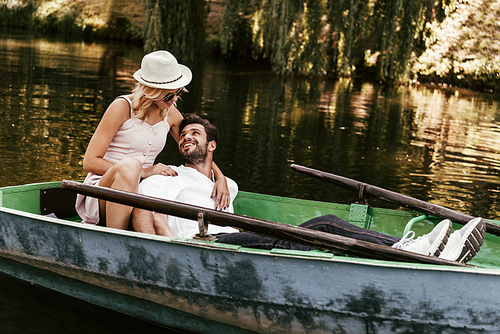 young woman in sundress and hat hugging happy boyfriend while sitting in boat