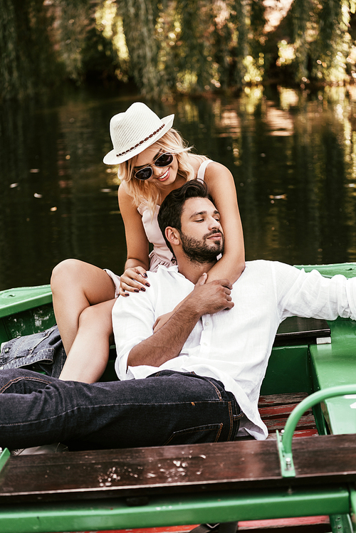 young woman in hat and sunglasses hugging handsome boyfriend while sitting in boat