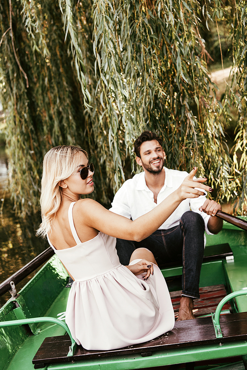 attractive young woman pointing with finger while sitting in boat near happy boyfriend