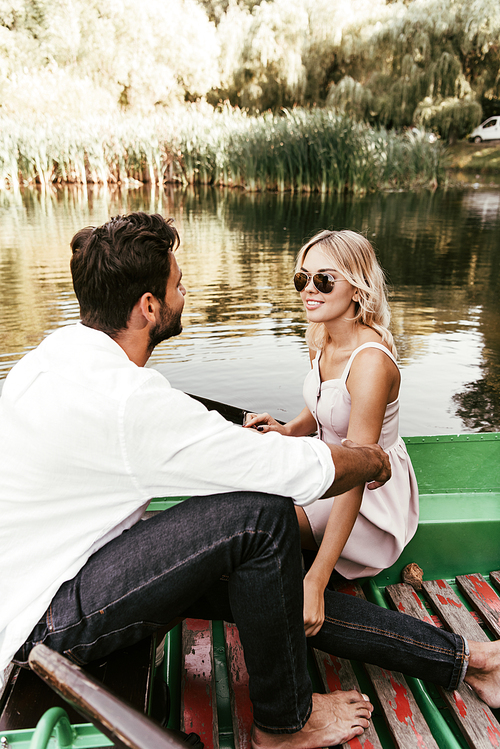 attractive, smiling woman looking at boyfriend while sitting in boat on lake