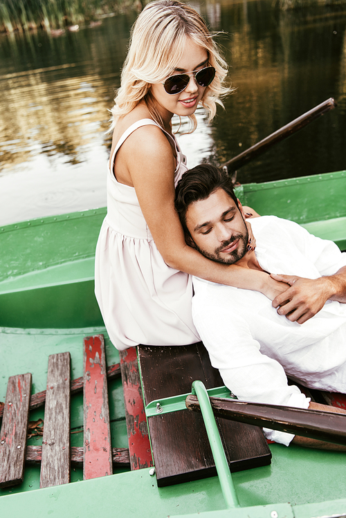 attractive woman in sunglasses hugging boyfriend while sitting in boat on lake