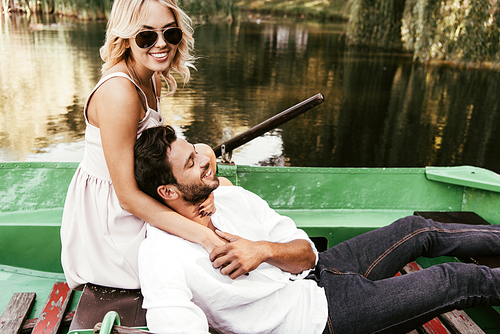 attractive girl in sunglasses hugging happy boyfriend while sitting in boat on lake