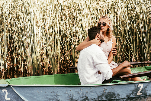 attractive young woman embracing boyfriend in boat on lake near thicket of sedge