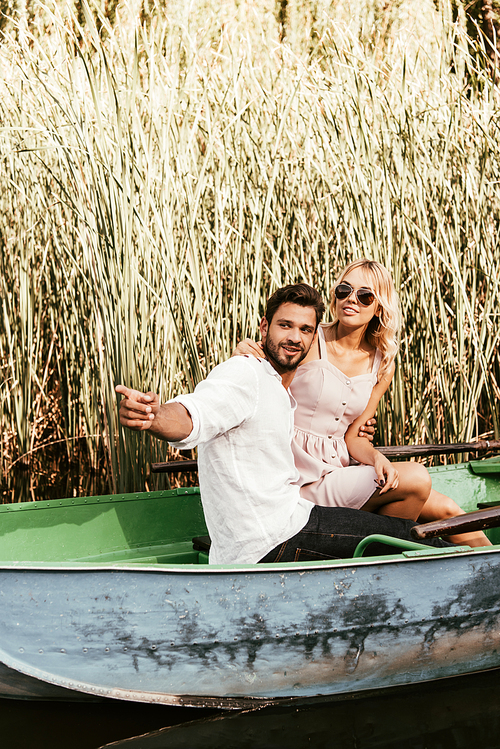 smiling man pointing with finger while sitting with girlfriend in boat near thicket of sedge