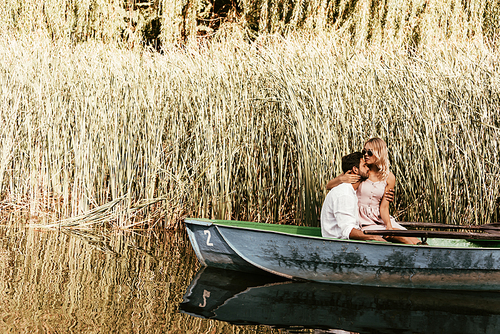 happy young couple embracing while sitting in boat on river near thicket of sedge