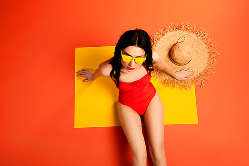 top view of young woman in sunglasses and swimsuit sitting near straw hat on orange