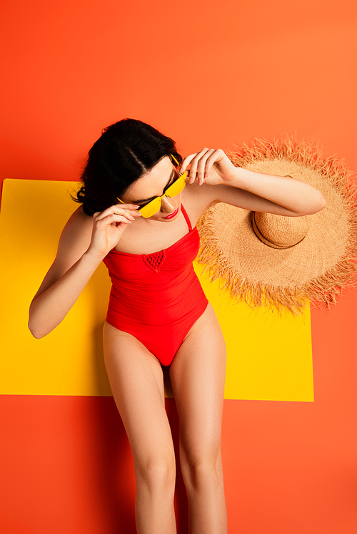 top view of young woman in swimsuit touching sunglasses while sitting near straw hat on orange