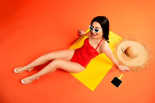 top view of cheerful woman in sunglasses and swimsuit sitting near straw hat and mirror on orange