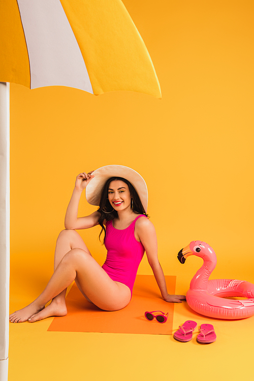 happy woman in straw hat and swimsuit sitting near sunglasses, flip flops, inflatable ring and paper umbrella on yellow