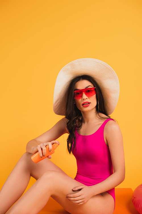 woman in straw hat, sunglasses and bathing suit holding bottle with sunscreen on yellow