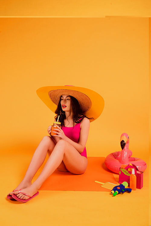 young woman in straw hat and bathing suit holding glass with orange juice near inflatable ring and water gun on yellow