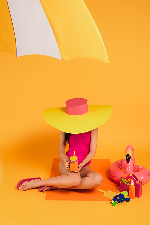 girl in straw hat and pink swimsuit holding glass with orange juice near water gun and inflatable ring on yellow