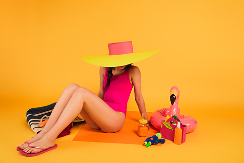 stylish woman in straw hat and pink swimsuit sitting near glass with orange juice, water gun and inflatable ring on yellow