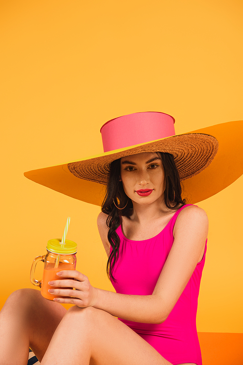 pretty girl in straw hat and swimsuit holding glass with orange juice on yellow